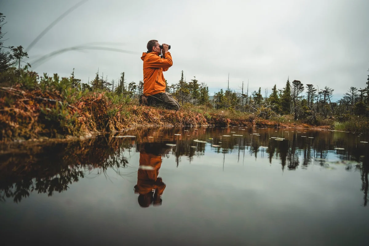 Man in orange jacket observing landscape with binoculars beside calm water, surrounded by forest and wetland plants.