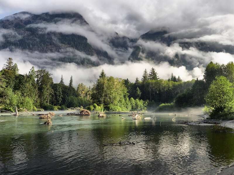 River surrounded by mist-shrouded mountains, green forest shoreline, and emerald water under cloudy skies.