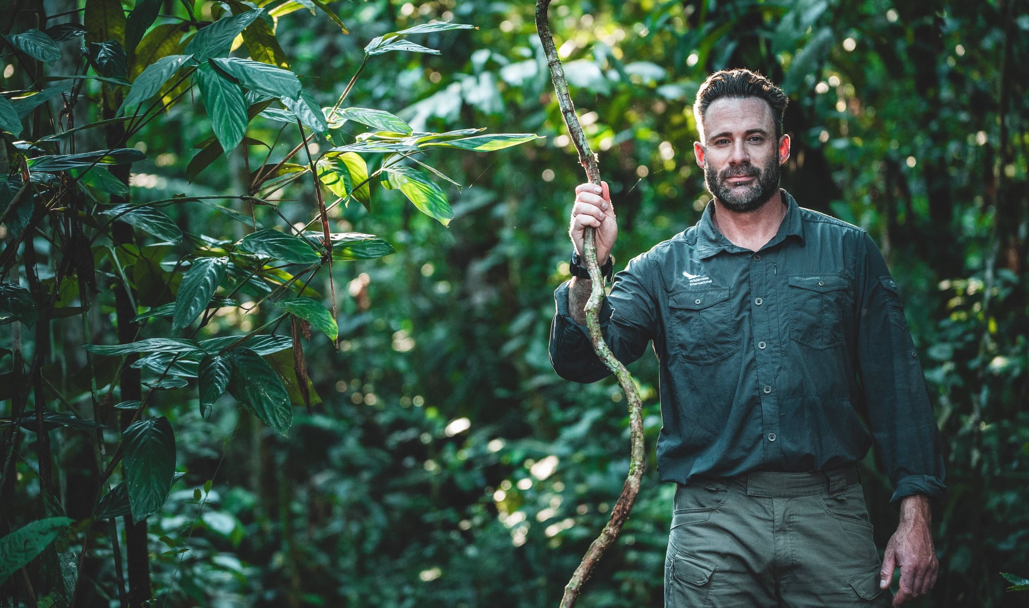Hombre con camisa oscura sosteniendo una rama de enredadera en la selva verde exuberante.
