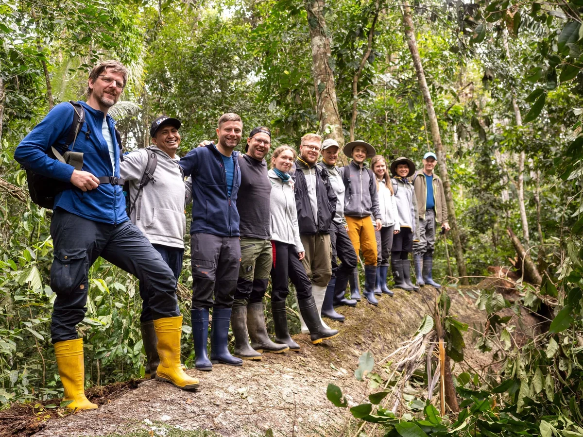 Group of about twelve people smiling on a forest trail surrounded by lush green trees and vegetation.