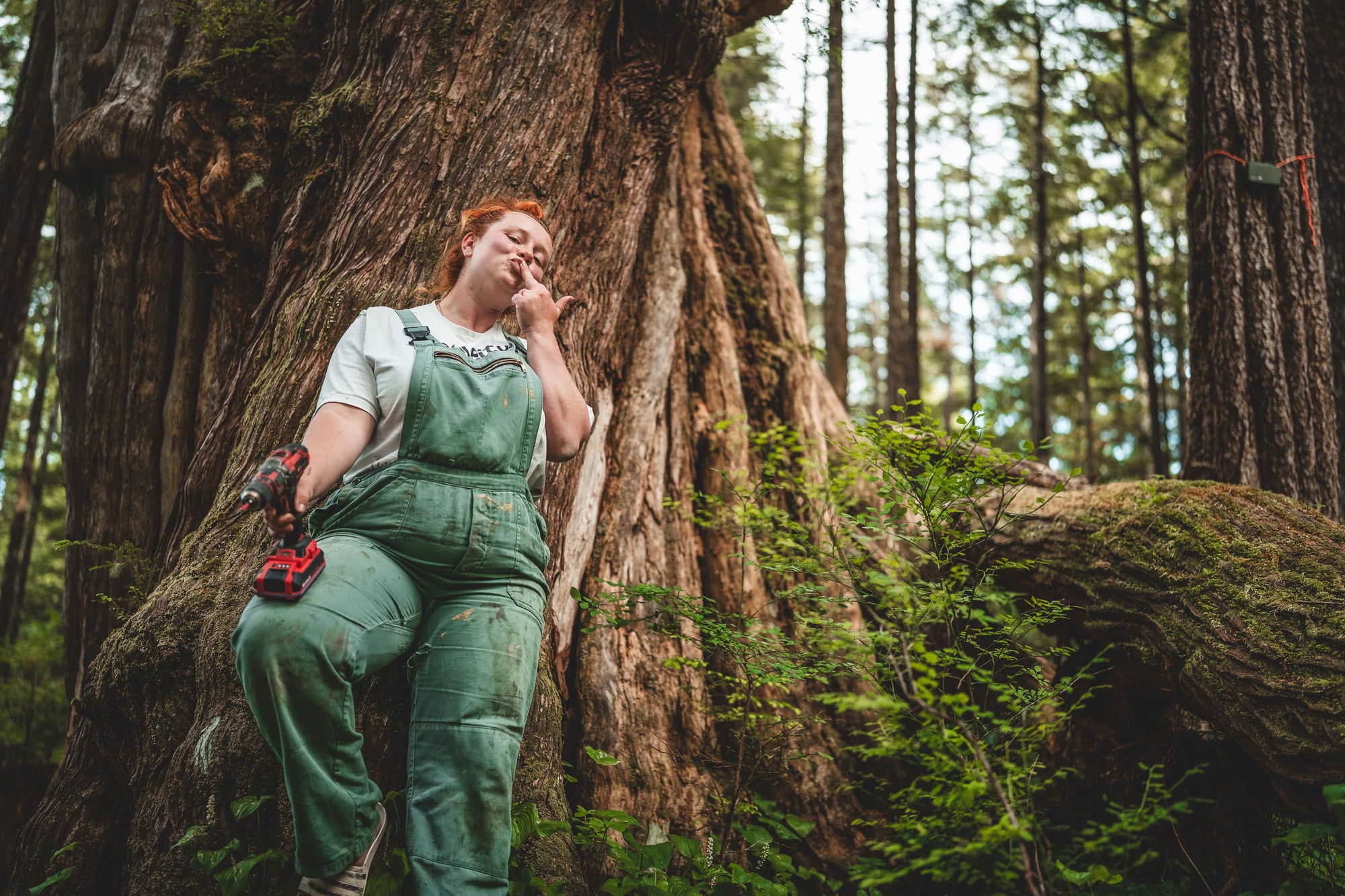 Woman in green work overalls leans against a massive tree trunk in the forest, holding a red chainsaw.