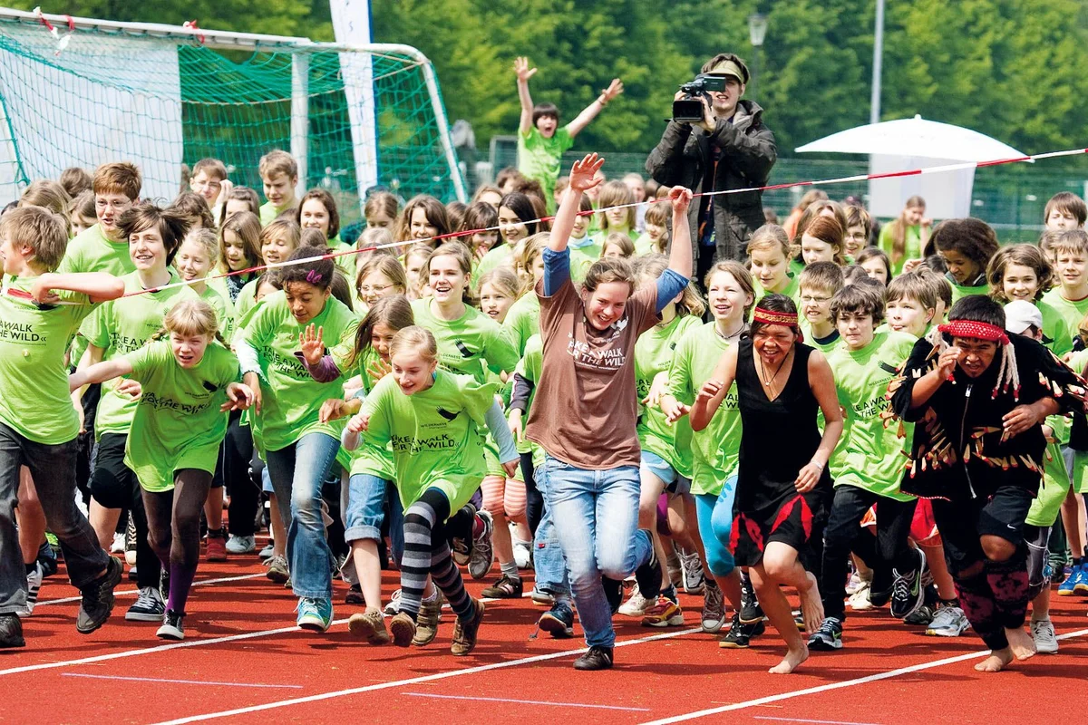 Grupo numeroso de niños con camisetas verdes corriendo en una pista roja durante un evento deportivo.