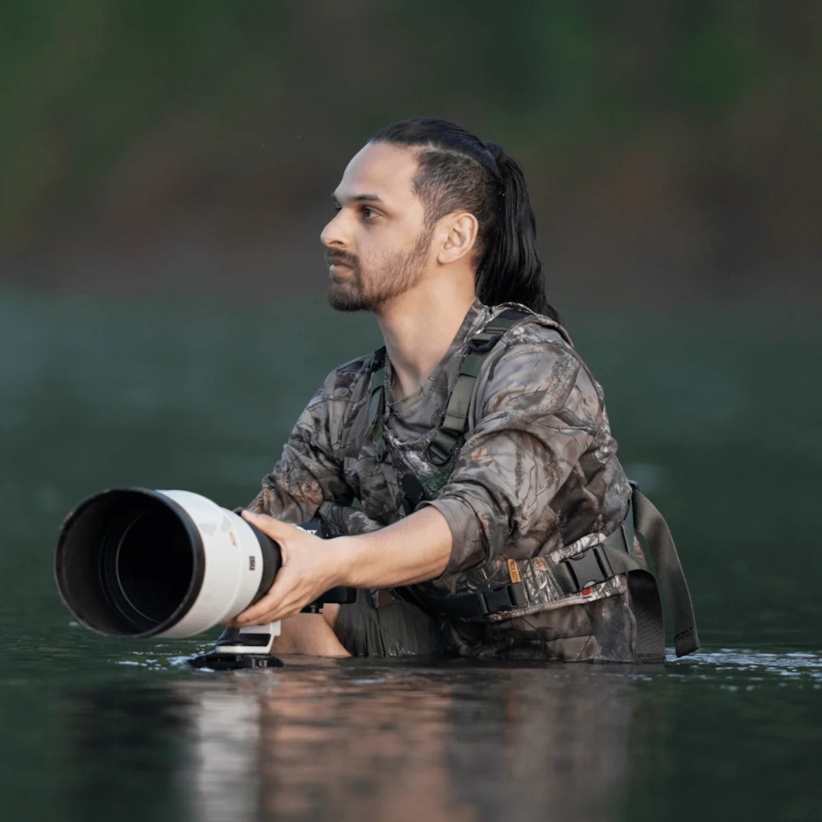 Hombre con cabello largo oscuro y barba arrodillado en el agua, sosteniendo una lente teleobjetivo grande.