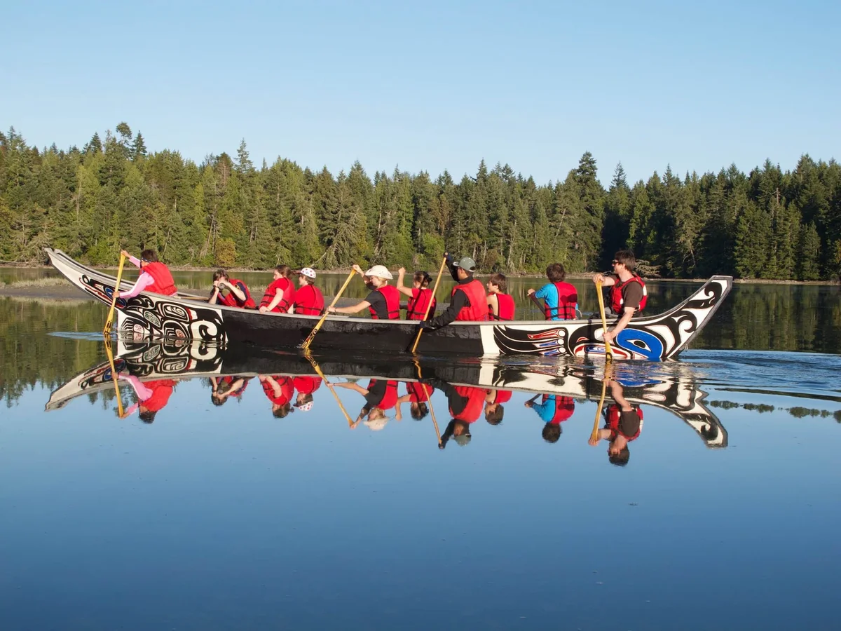 Group paddling an ornately carved canoe in calm water, wearing red, with coniferous forest backdrop.