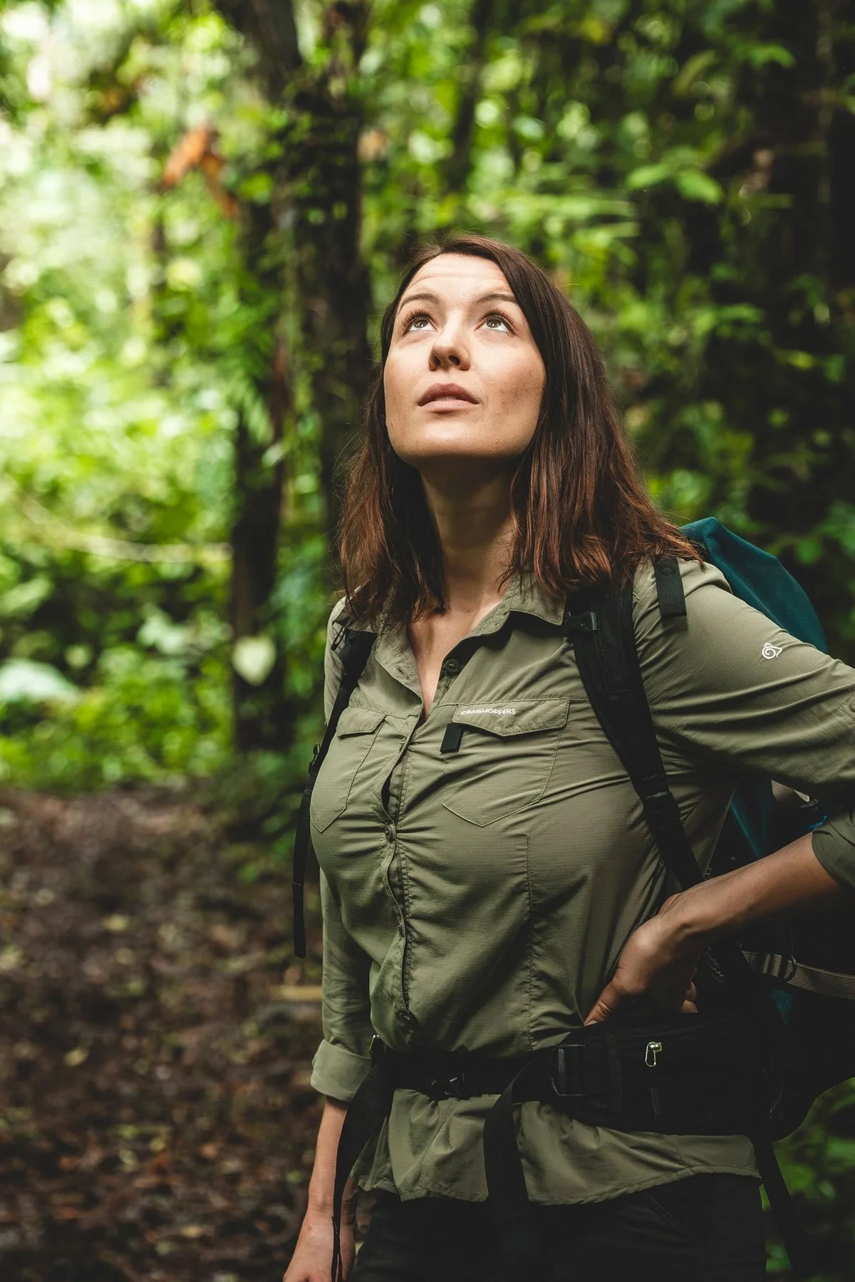 Mujer de cabello castaño con camisa caqui y mochila mirando hacia arriba en un bosque verde exuberante.