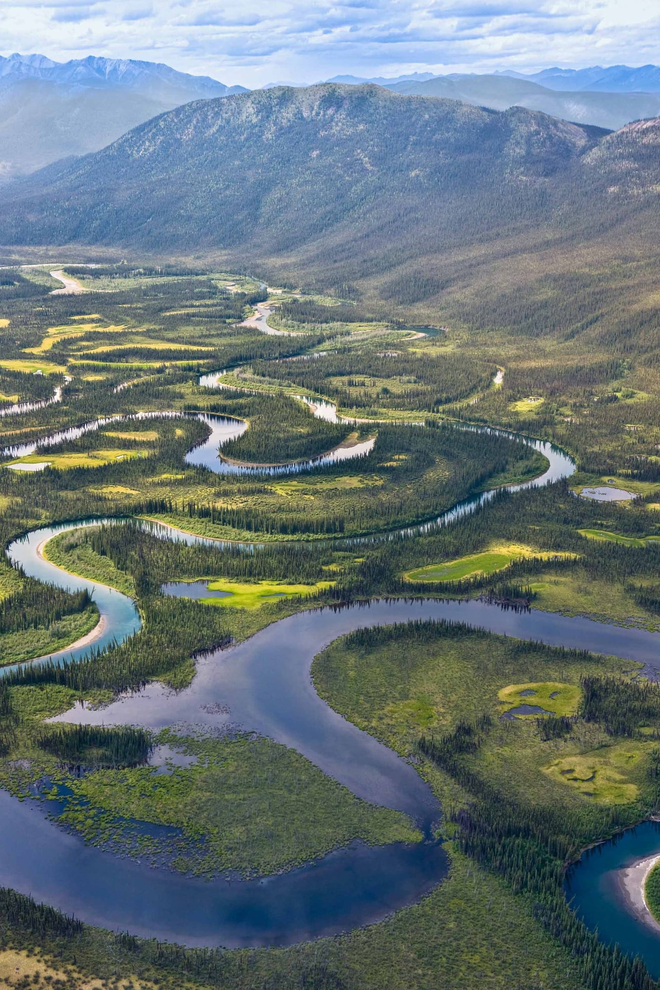 Aerial view of a meandering river valley surrounded by boreal forest, wetlands, and snow-capped mountains.