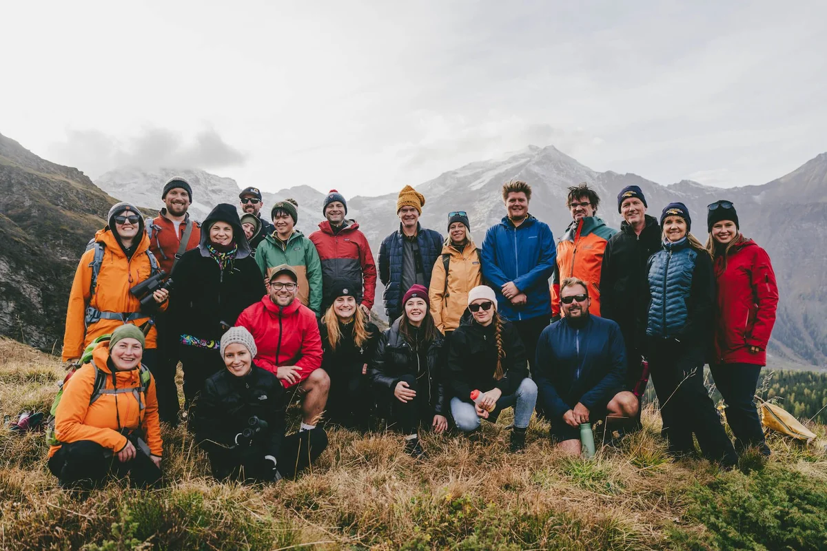 Large hiking group in colorful jackets posing in front of snow-capped alpine mountains and green valley.