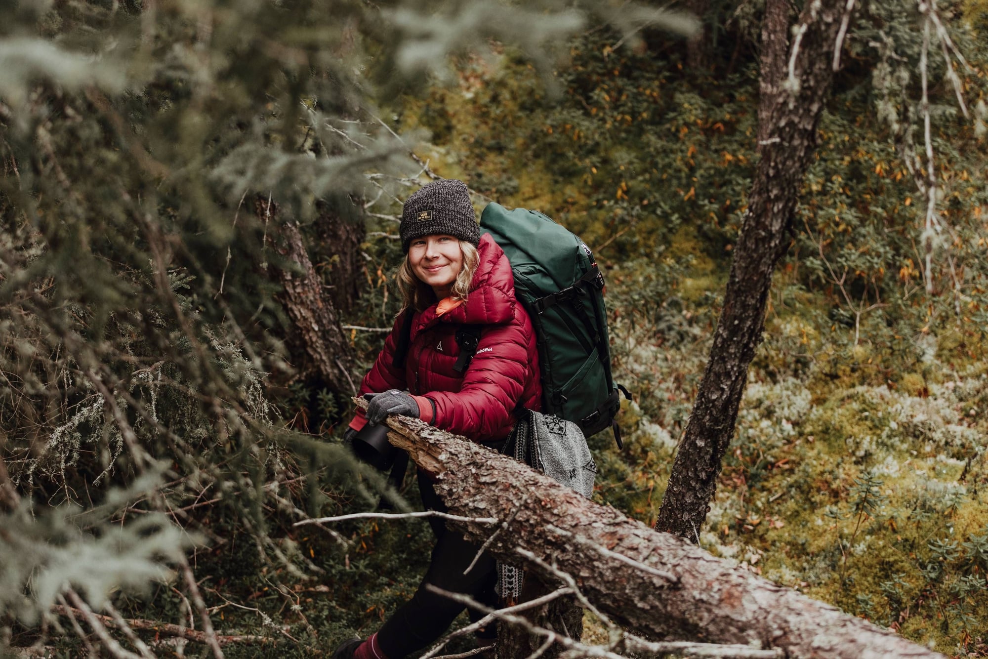 Excursionista sonriente con chaqueta roja, gorro gris y mochila se sienta sobre un tronco caído en el bosque.