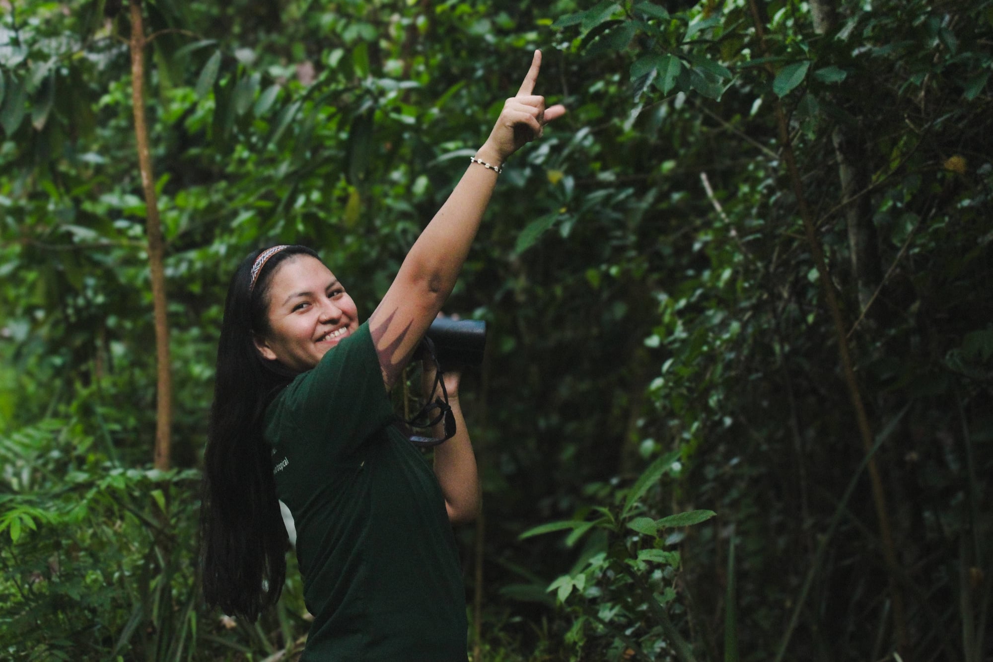 Smiling woman with long dark hair pointing upward in a lush green forest setting.
