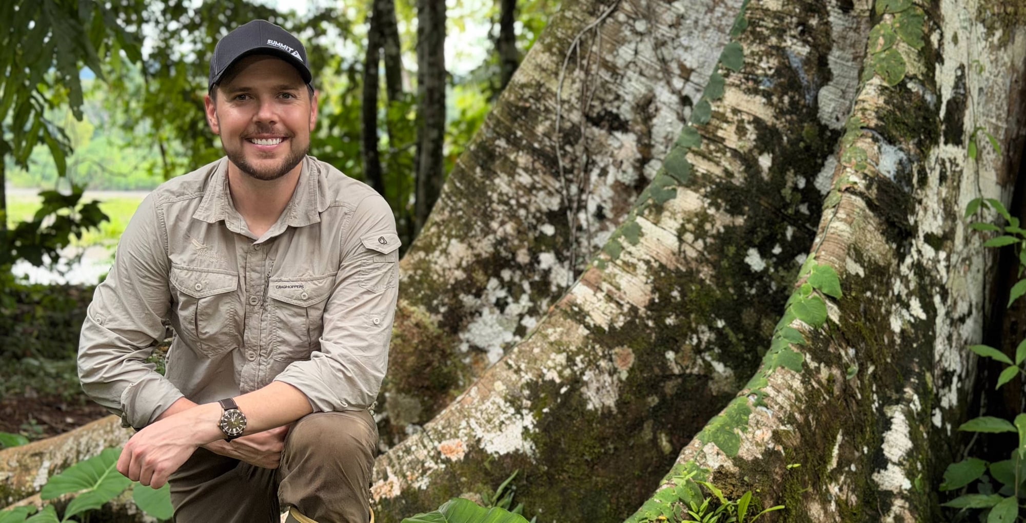 Smiling man wearing khaki shirt and baseball cap crouches beside moss-covered tree trunks in forest setting.