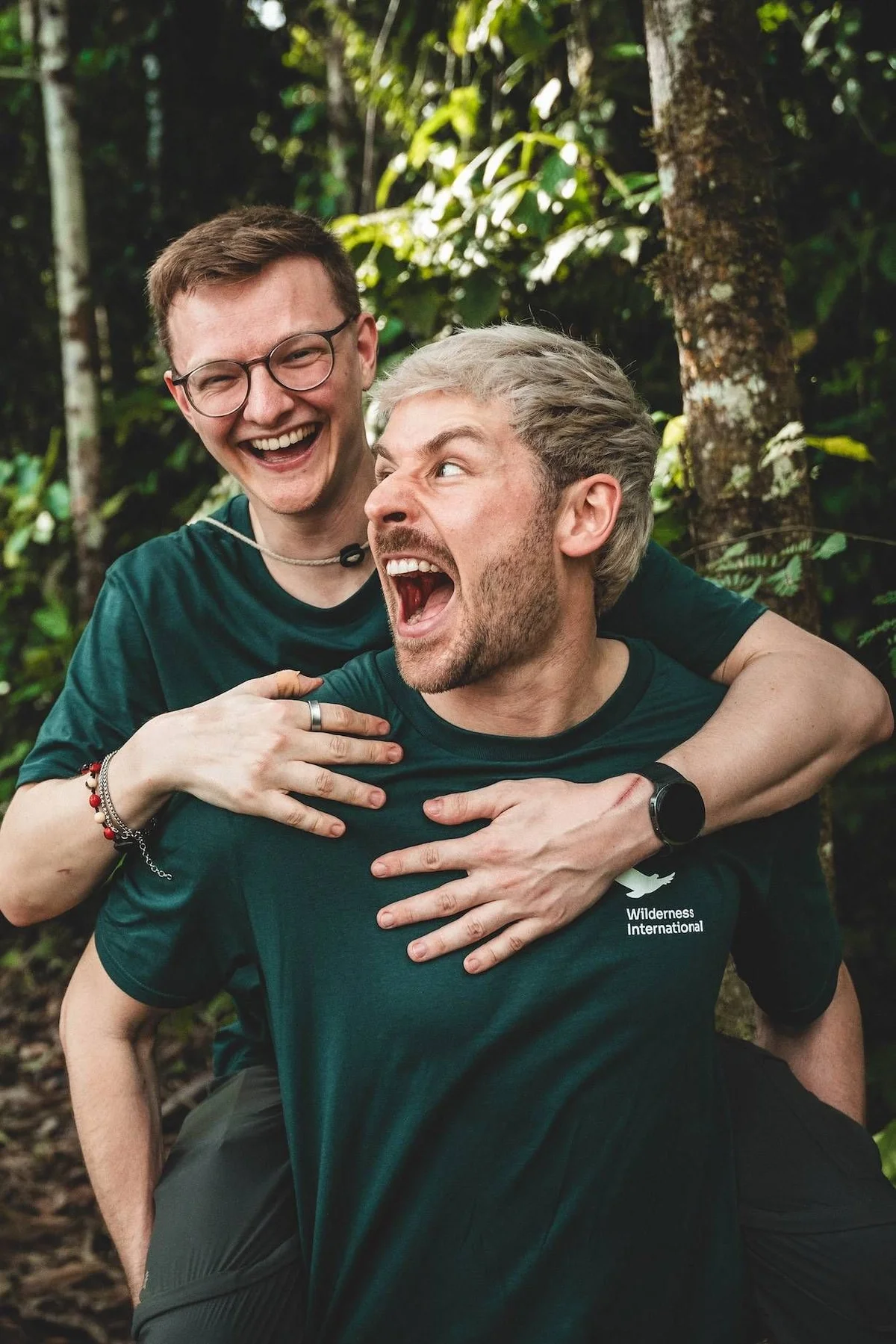 Dos hombres riendo abrazo en el bosque usando camisetas verdes de Wilderness International, con árboles y vegetación exuberante de fondo.