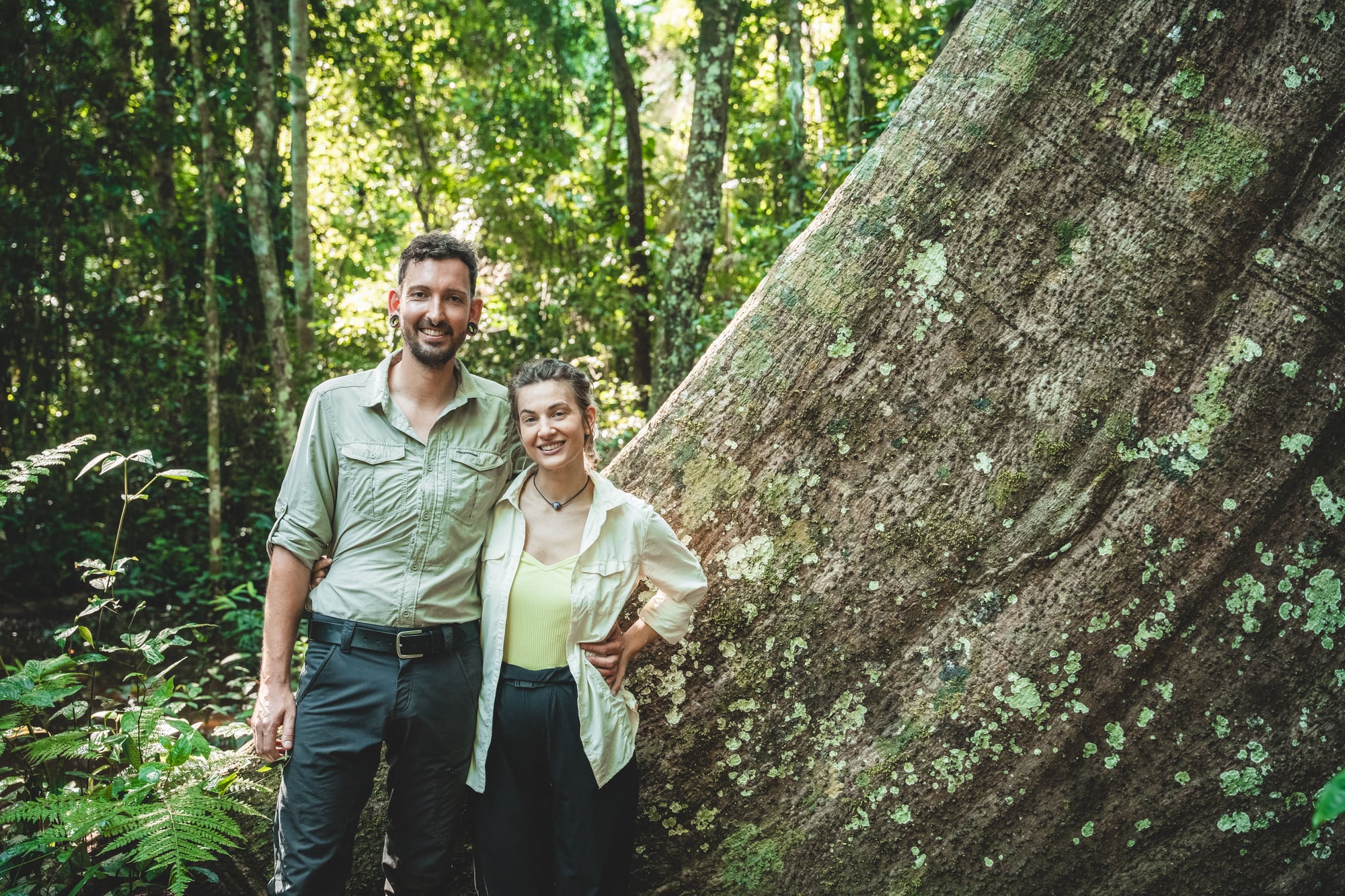 Un hombre y una mujer sonríen junto a un árbol gigante cubierto de musgo en un bosque exuberante.