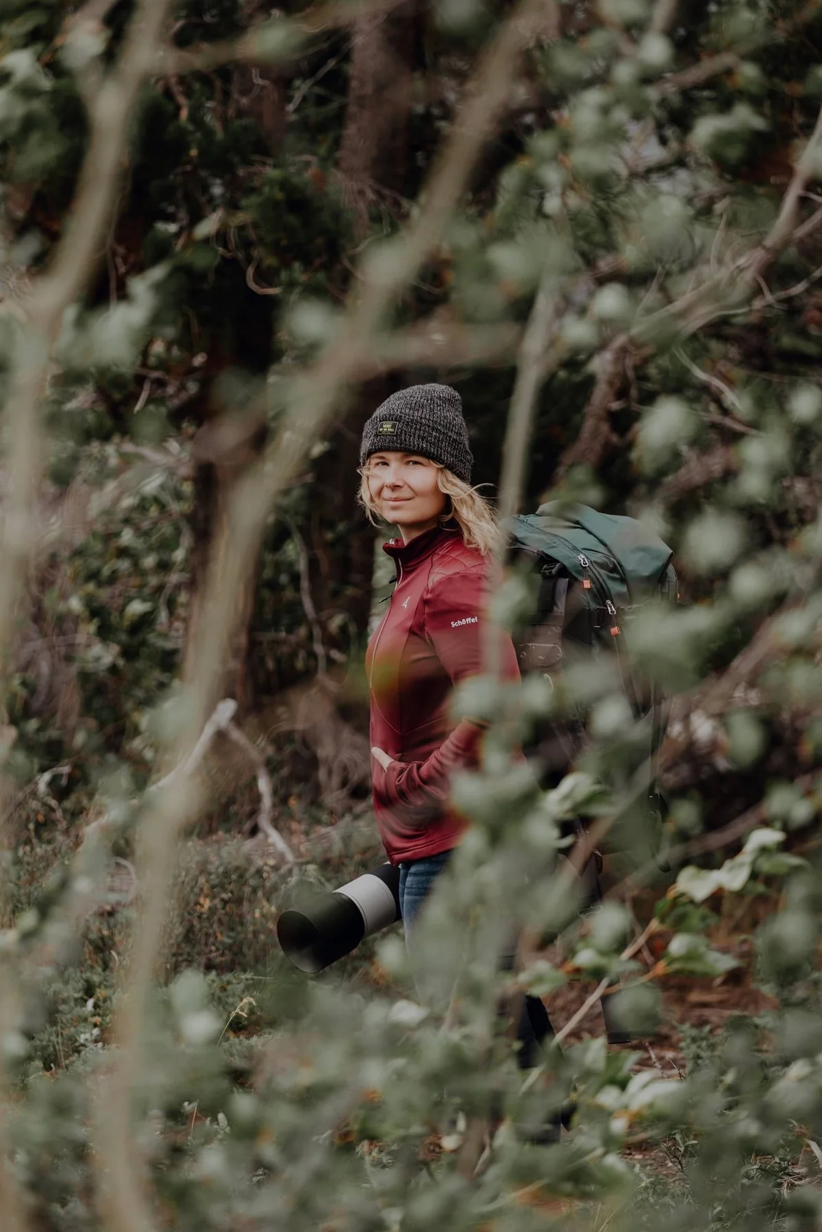 Mujer con gorro gris y chaqueta roja caminando entre arbustos y árboles