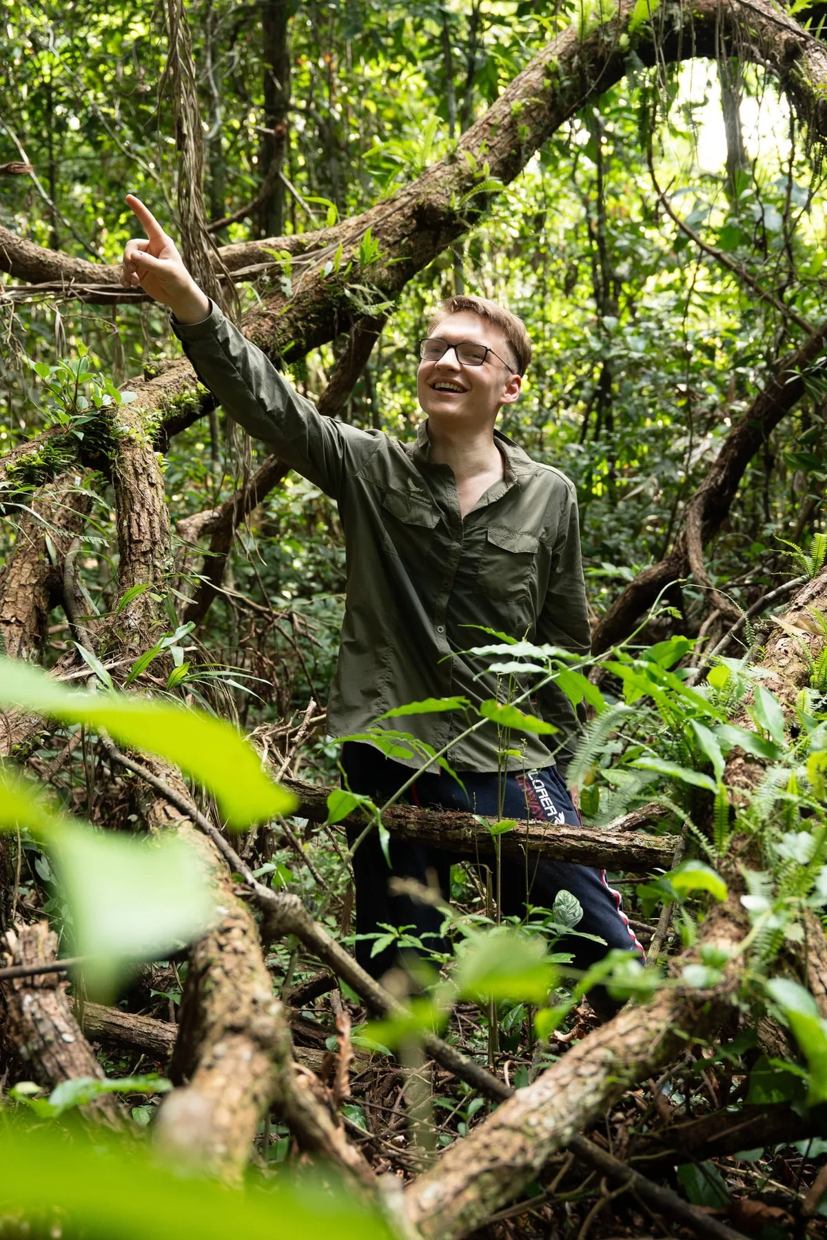 Young man wearing glasses explores lush jungle vegetation, surrounded by fallen logs and vibrant green foliage.