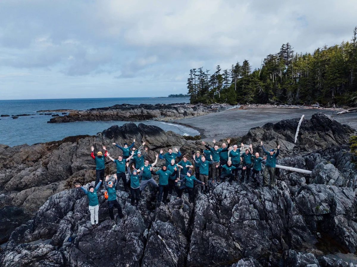 Large group wearing teal shirts posing on rocky coastal rocks with ocean, forest, and cloudy sky in background.
